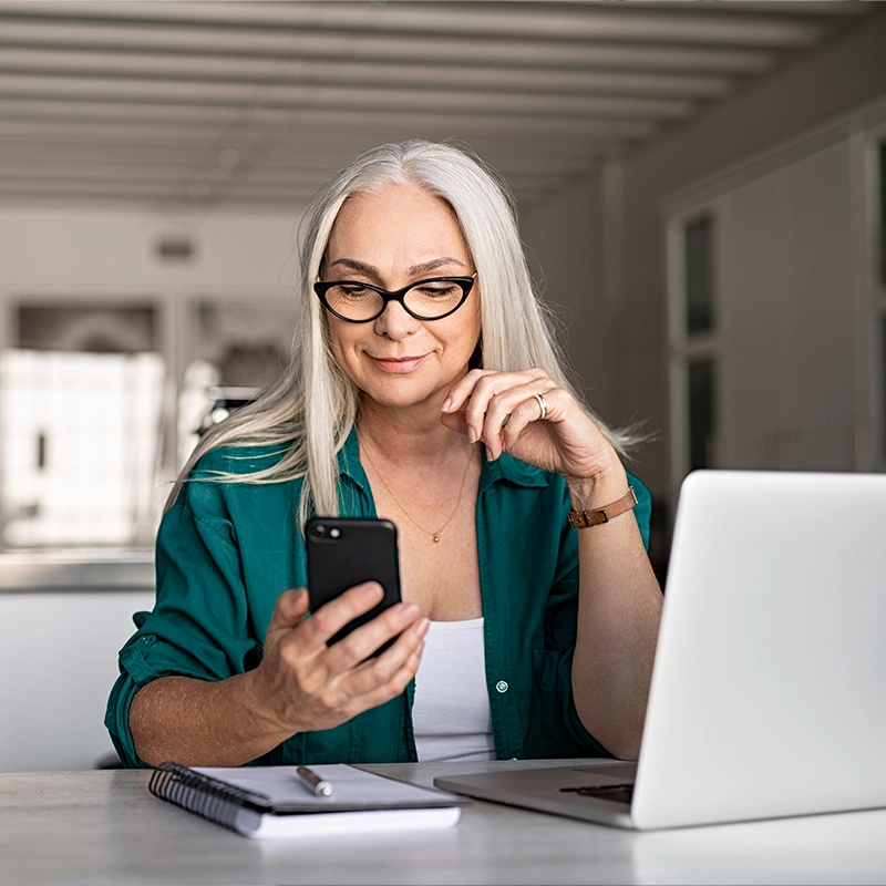 image of young mature woman smiling using cell phone and laptop computer in home