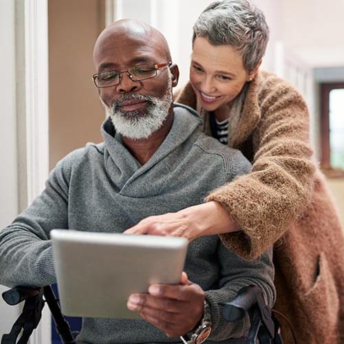 A woman leaning over a man in a wheelchair pointing at the tablet he his holding